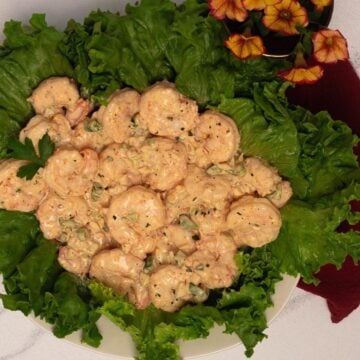 Shrimp remoulade on top of green leaf lettuce on a white plate next to a dark red napkin and yellow and red flowers.
