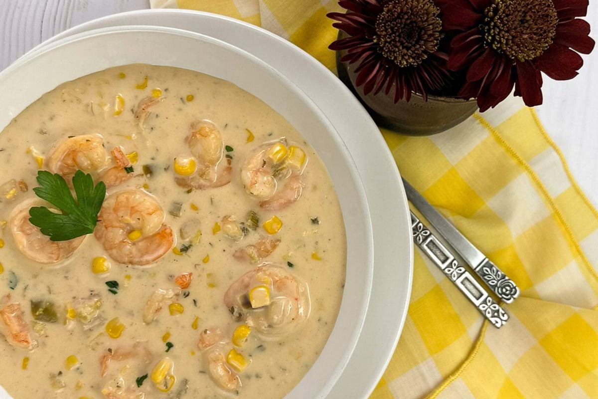 Shrimp and corn bisque in a round white bowl with a yellow and white napkin and dark red flowers. 
