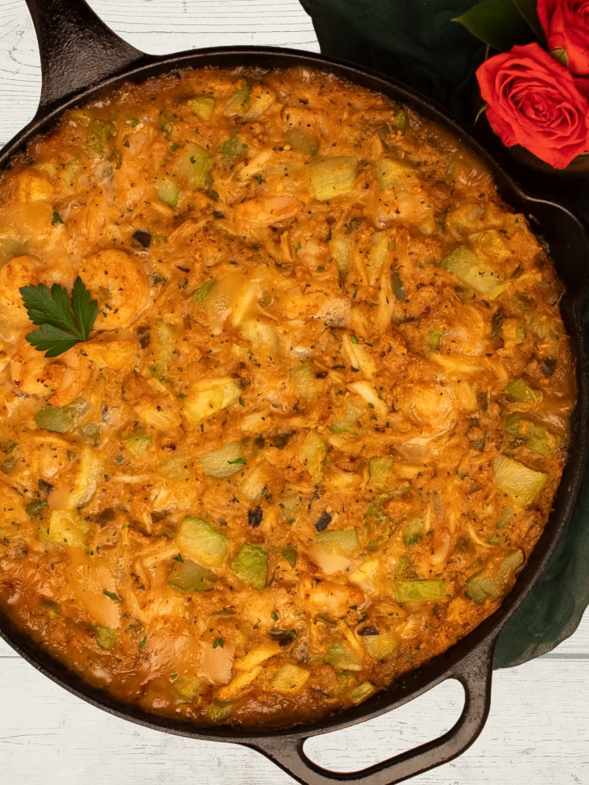 Mirliton dressing with shrimp and crab meat in a black cast iron skillet alongside orange roses and a dark green napkin.