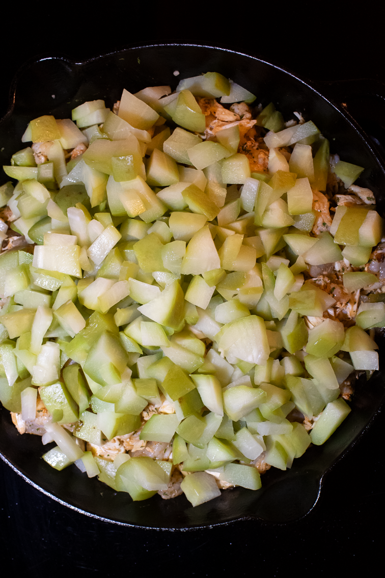Chopped cooked mirliton squash on top of vegetables and seafood in a pan.