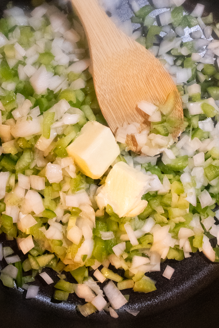 Chopped onion, bell pepper, celery, butter and oil with a wooden spoon in a black cast iron skillet.