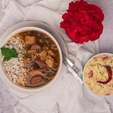 Chicken and sausage gumbo on a light background with a white napkin, potato salad side, and red flowers.