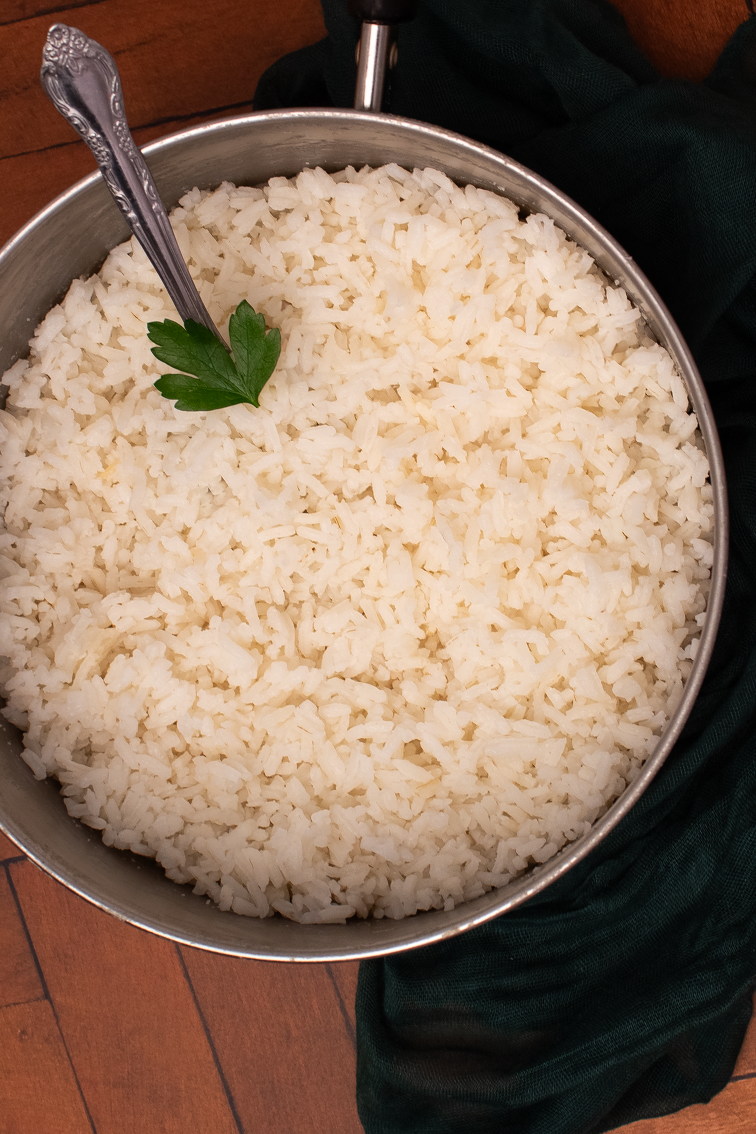Cooked white rice in a small pot with a whole parsley leaf garnish and a spoon handle in the top left side of the pot.