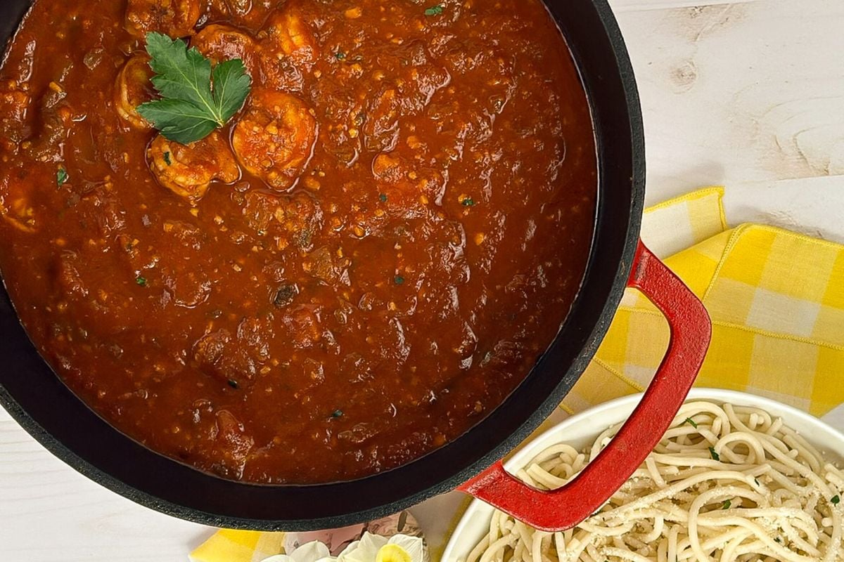 Cajun shrimp spaghetti in a red enameled cast iron Dutch oven along spaghetti noodles.