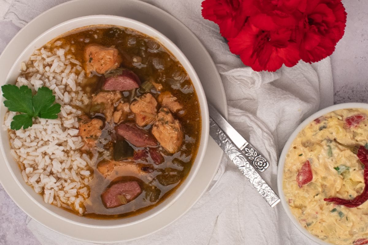 Chicken and sausage gumbo in a round white bowl on a white saucer with a napkin, side of potato salad, and red flowers.