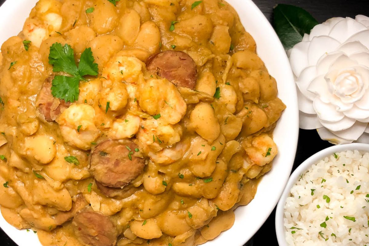 Cajun butter beans with shrimp and sausage on a round white bowl alongside rice and a flower.