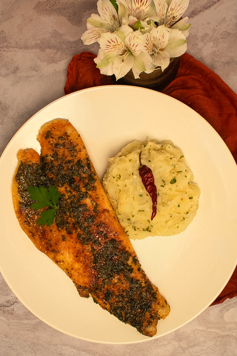 One trout meuniere fillet alongside parsley potatoes with a whole cayenne pepper garnish on a round white plate.