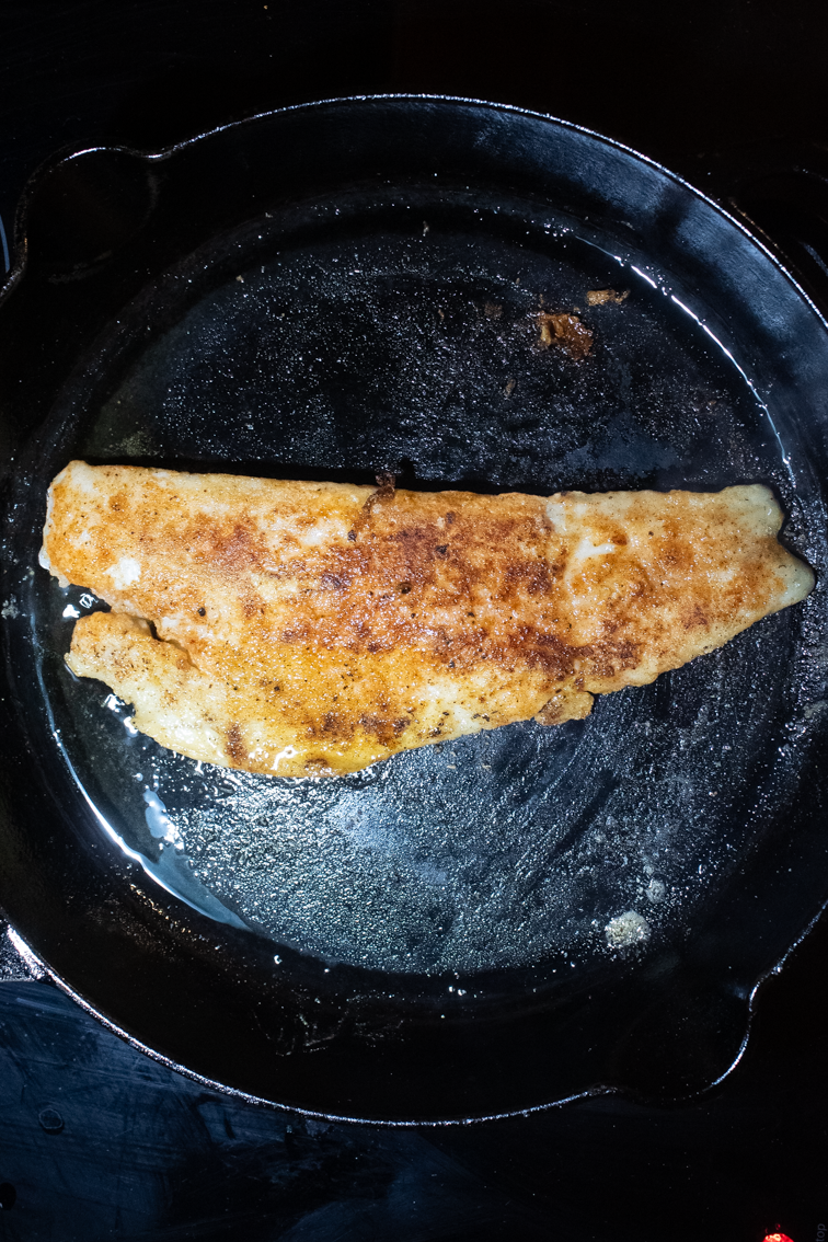 The browned side of a fillet for trout meunière in a black pan.