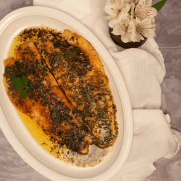 Two trout meunière fillets on an oval white serving platter alongside a white napkin and white flowers.