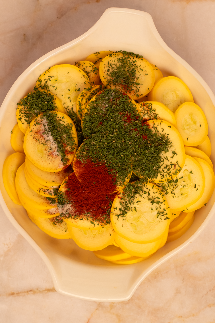 Sliced fresh yellow squash  with dried spices on top in a large white bowl. 