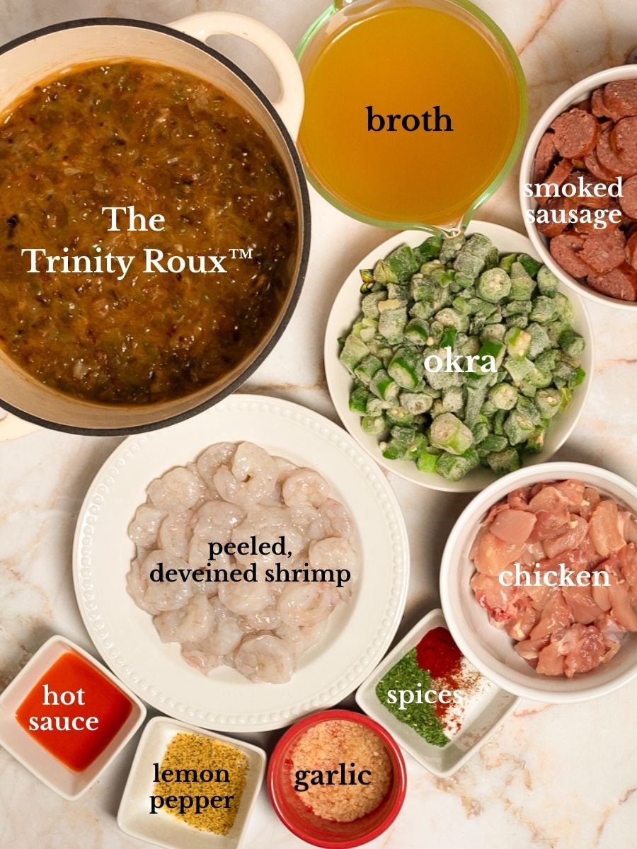 Labeled ingredients for chicken, sausage, and shrimp gumbo in bowls on a white marbled background.
