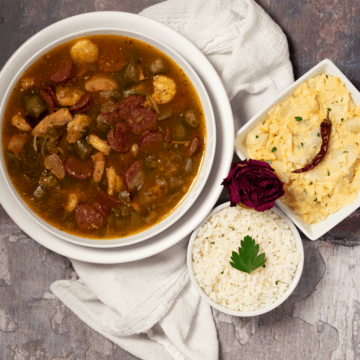 Chicken, sausage, and shrimp gumbo alongside a white napkin, dried red rose, small bowl of rice and side of potato salad.