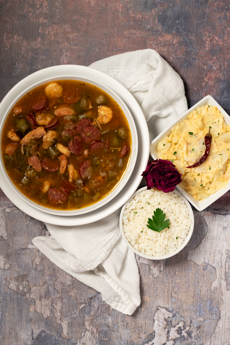 Chicken, sausage, and shrimp gumbo alongside a white napkin, dried red rose, small bowl of rice and side of potato salad.