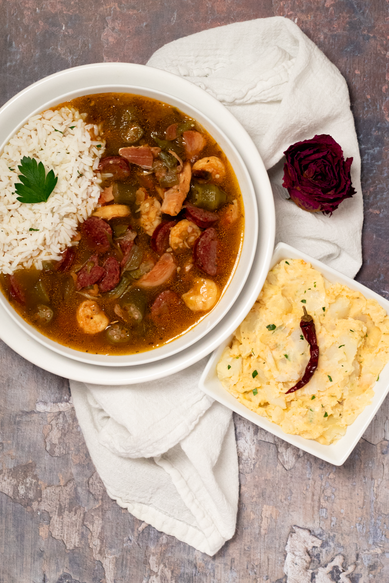 Chicken, sausage, and shrimp gumbo in a round white bowl alongside a side of potato salad.