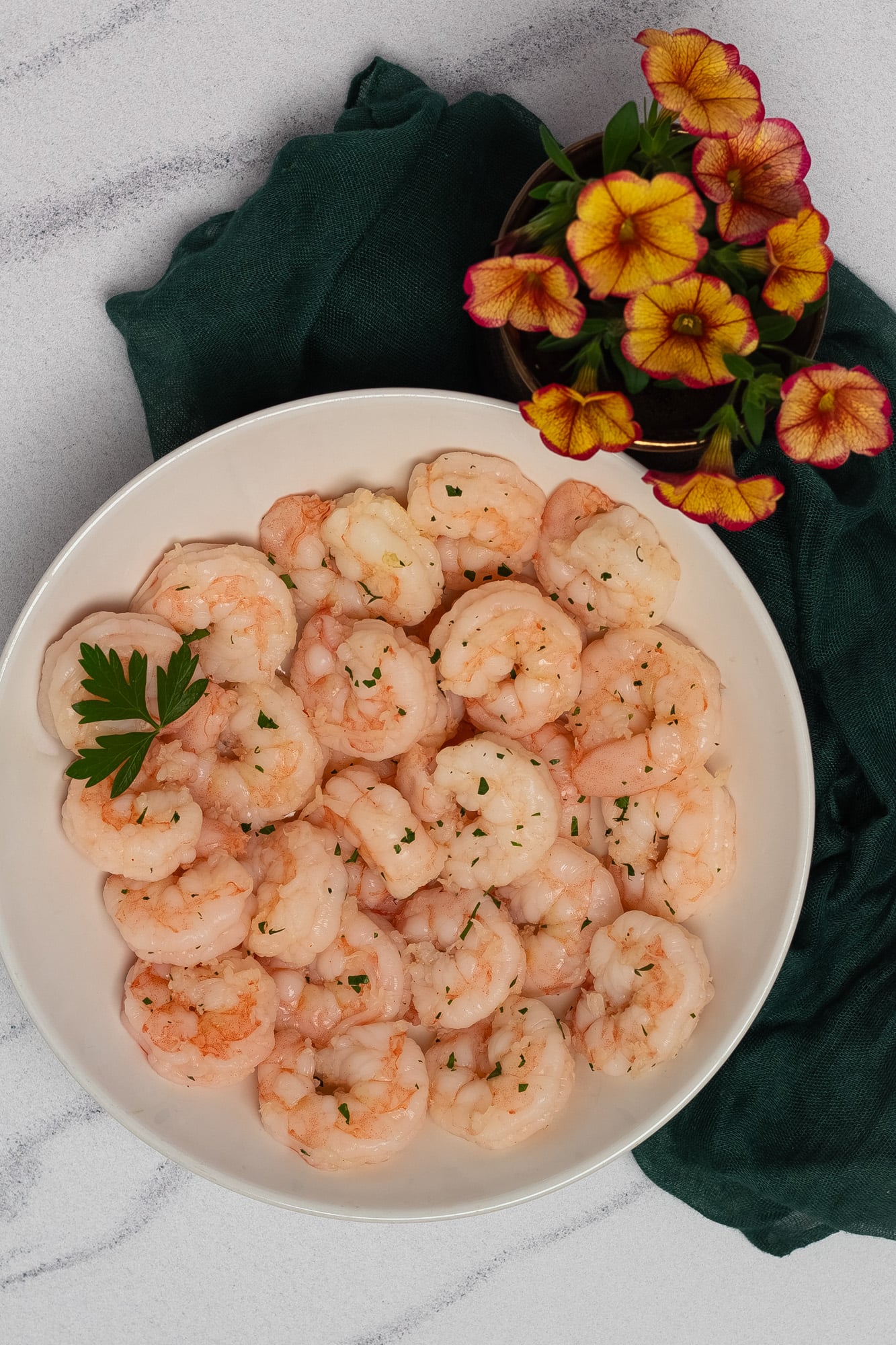 A round white bowl filled with poached shrimp alongside a dark green napkin and yellow and red flowers.