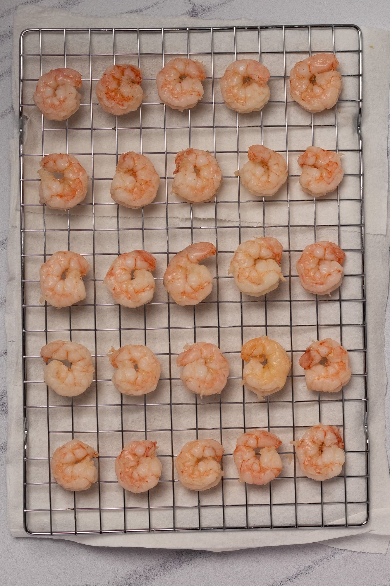 25 poached shrimp arranged in rows of 5 on a wire baking rack set atop a paper towel-lined aluminum baking sheet.