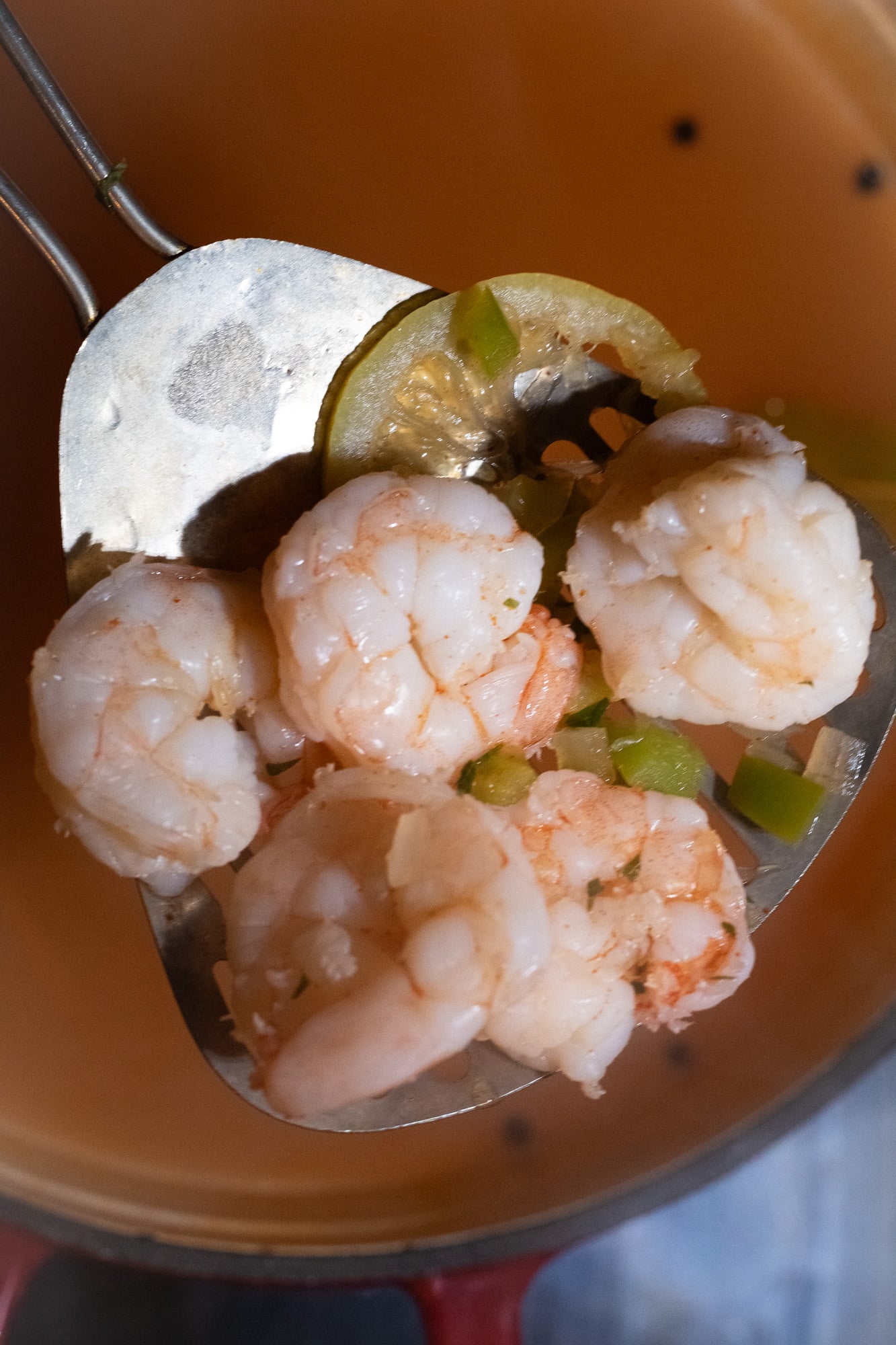 A slotted metal skimmer holding about 5 poached shrimp, a slice of lemon, and finely chopped green bell peppers, celery, and onions.