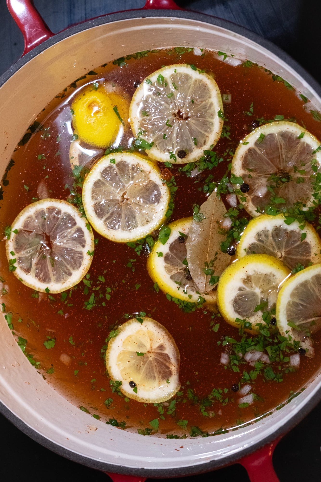 A poaching liquid featuring water, lemon slices, herbs, spices, and finely chopped seasoning veggies in a red enameled cast iron Dutch oven. 