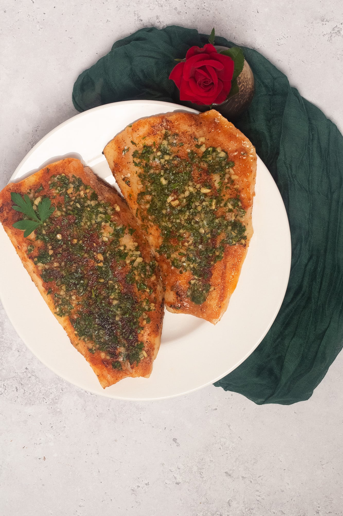 Two pan seared snapper fillets on a round white plate alongside a green linen napkin and a red rose.