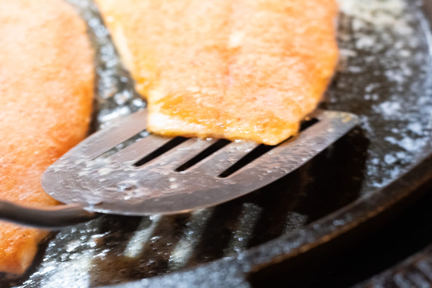 A wire spatula under a fish fillet cooking in a pan.