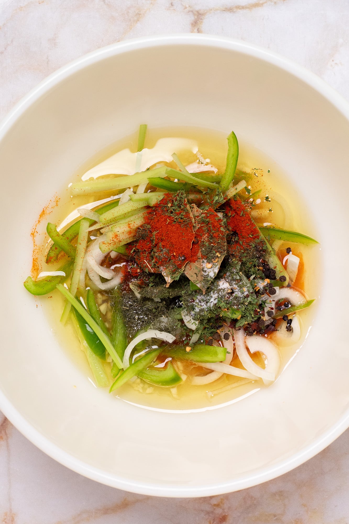 Avocado oil, lemon juice, finely sliced veggies, and dried spices in a large white vintage mixing bowl.