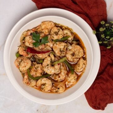 A round white bowl filled with pickled shrimp atop a round white saucer alongside a dark red napkin and green flowers.