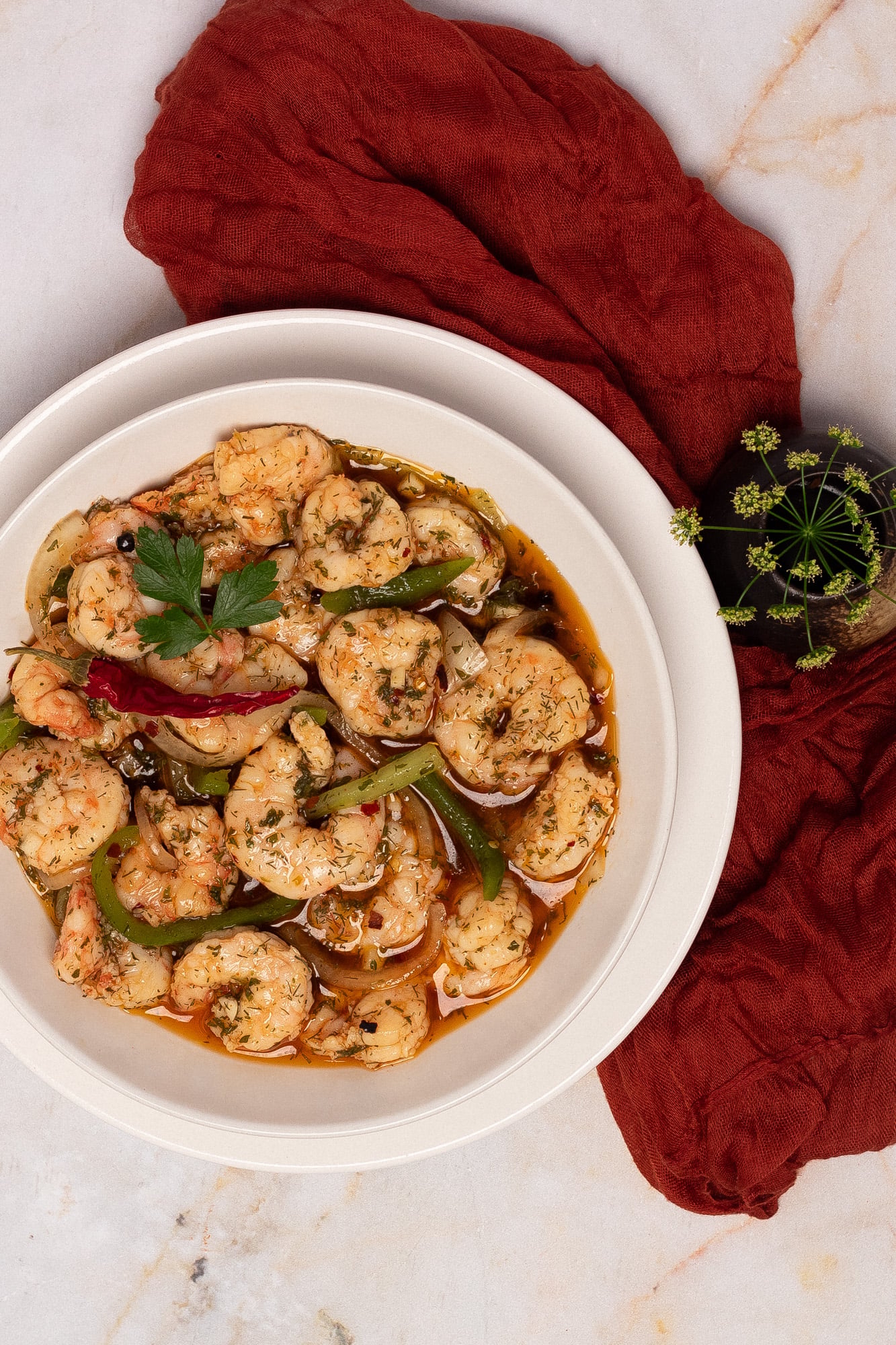 A round white bowl filled with pickled shrimp next to a dark red linen napkin and green flowers.