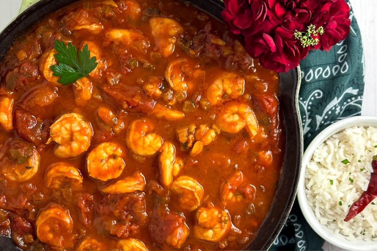 South Louisiana Shrimp Creole in a green enameled cast iron skillet alongside a green napkin and small bowl of rice topped with a cayenne pepper.