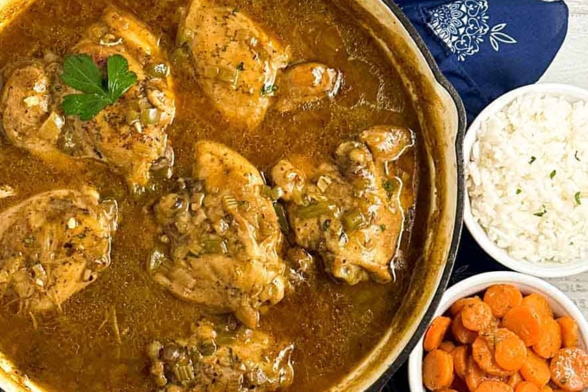 Cajun chicken fricassee in a red enameled cast iron skillet alongside rice, carrots, and a dark blue linen napkin.