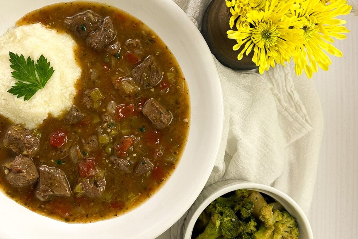 Grillades and grits in a small round white bowl alongside a side of broccoli and yellow flowers.
