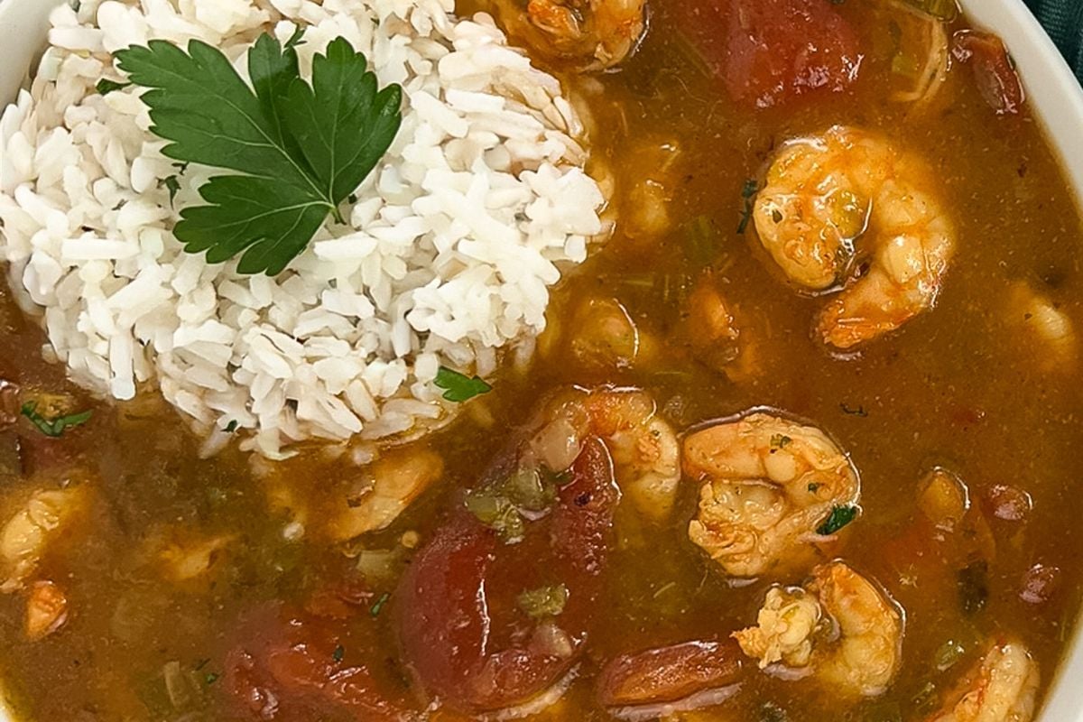 A closeup image of shrimp etouffee in a round white bowl with rice and a whole parsley leaf garnish on top.