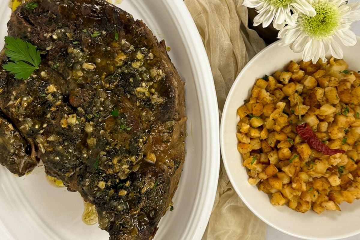 Louisiana garlic stuffed beef roast on a white plate alongside roasted potatoes and a bud vase with white flowers.