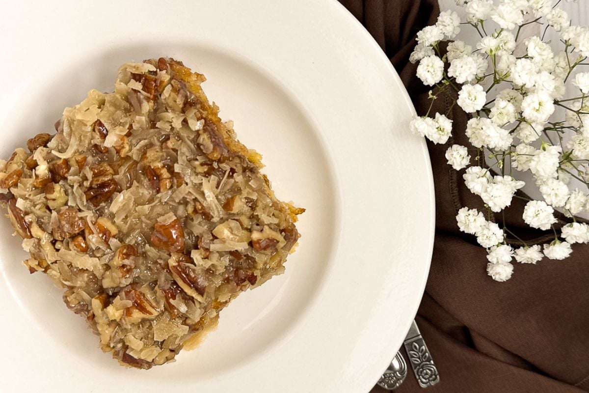 A slice of Cajun cake on a round white plate alongside baby's breath flowers in a vase and a brown napkin. 