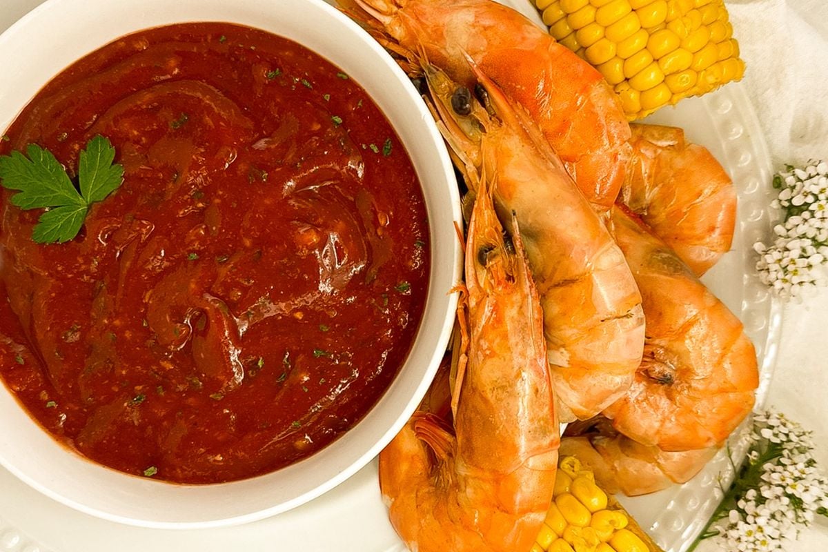 Cajun cocktail sauce in a white bowl alongside boiled shrimp and corn. 