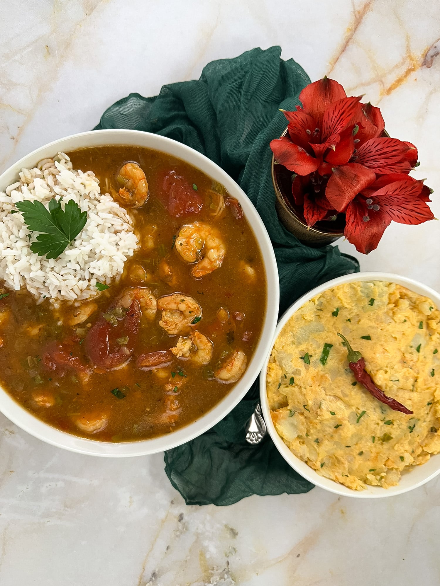 Shrimp etouffee in a round white bowl with rice on top alongside a green napkin, red flowers, and a bowl of potato salad.