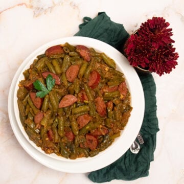 Cajun smothered green beans on a round white plate alongside a green linen napkin and dark red flowers.
