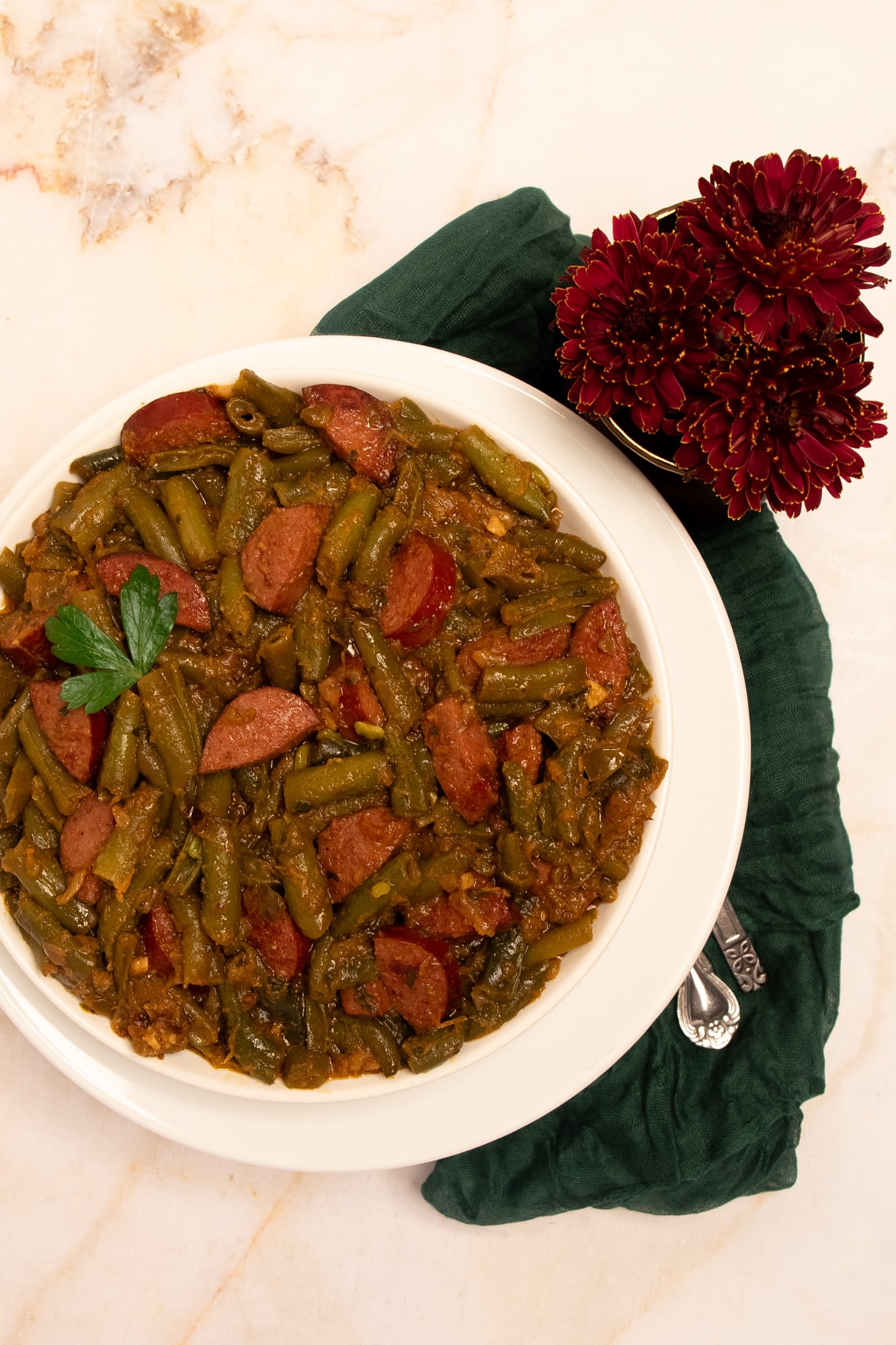 Cajun smothered green beans with sausage on a round white plate with a whole parsley leaf garnish alongside a green napkin and dark red flowers.