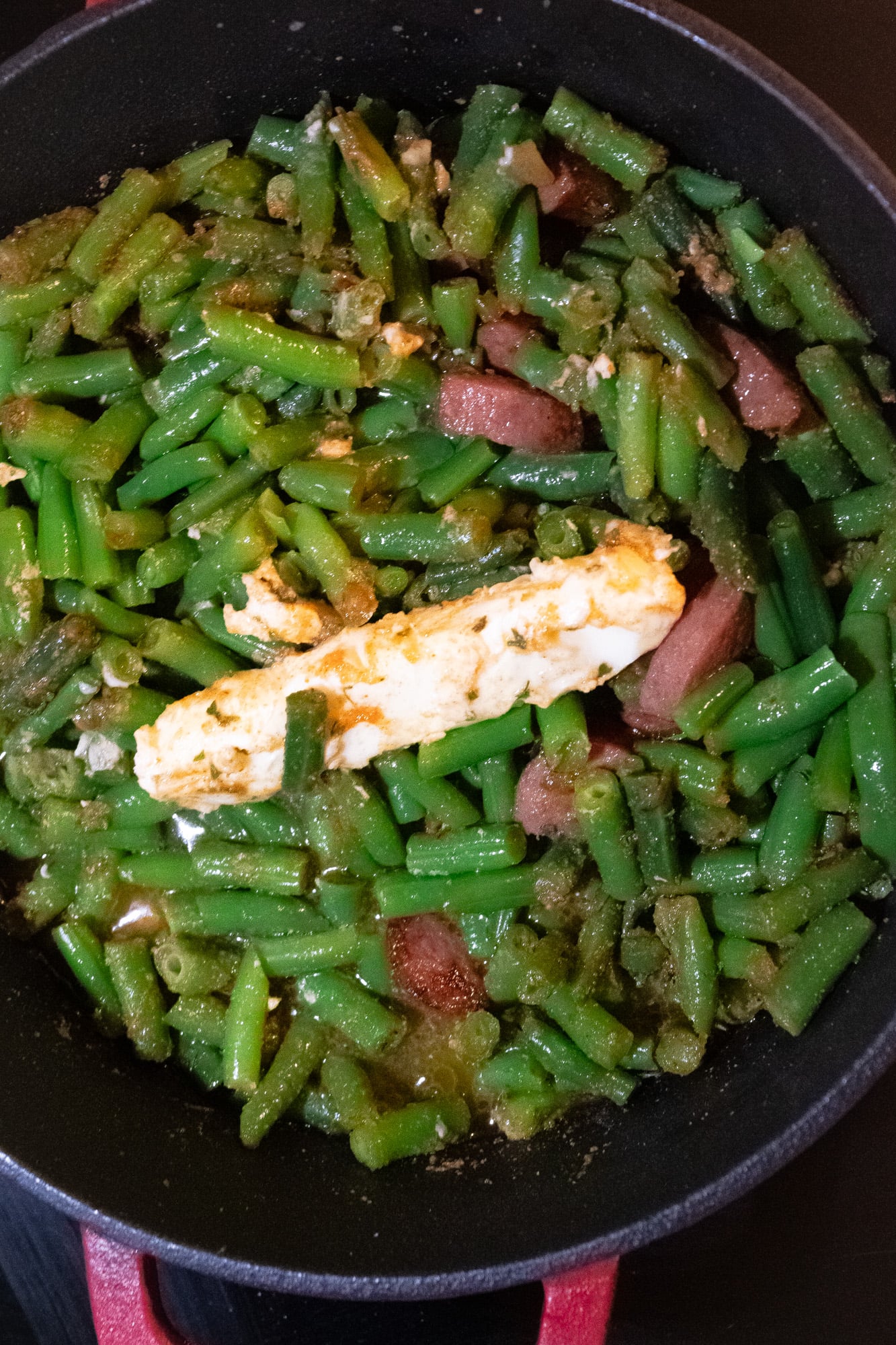 Seasoned green beans, butter, garlic, browned sausage, and browned vegetables in a red cast iron Dutch oven.